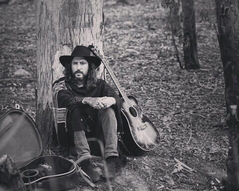 Paco Holguín con sombrero sentado en un bosque al lado de su guitarra