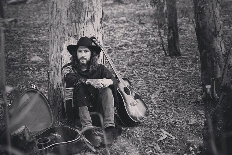 Paco Holguín con sombrero sentado en un bosque al lado de su guitarra