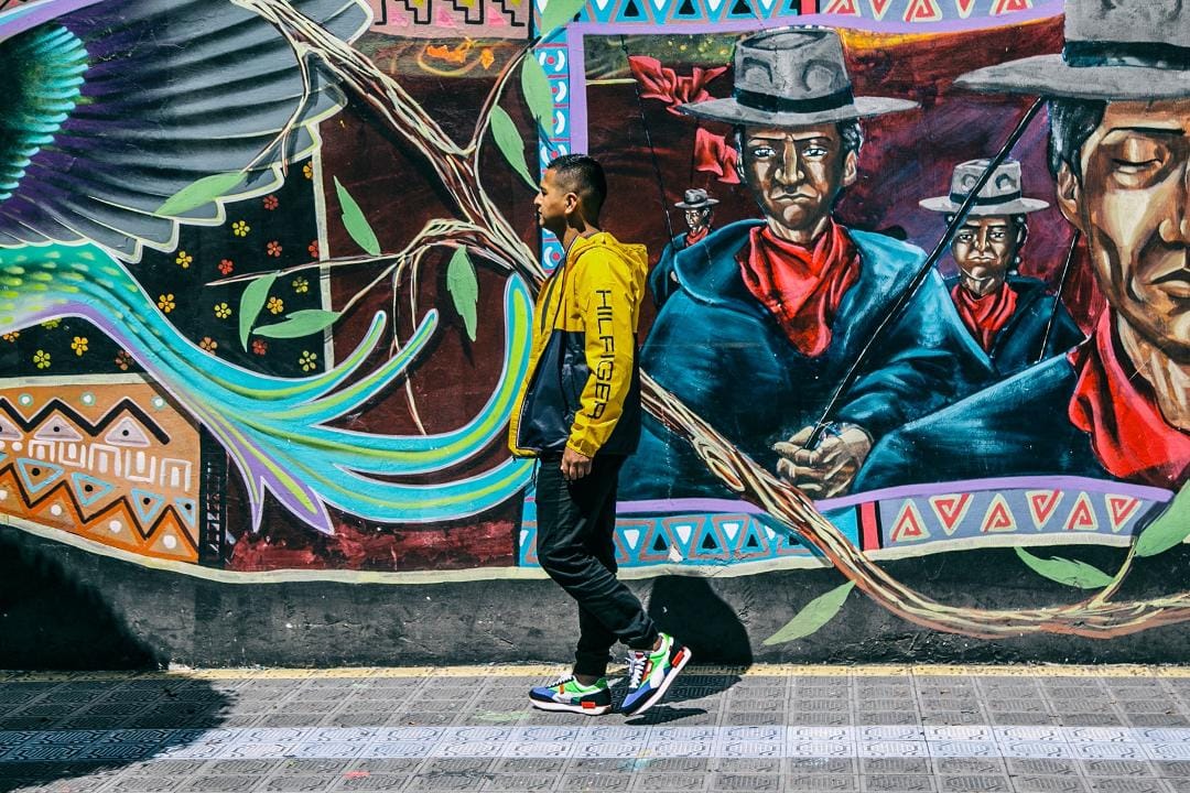 Liberato Kani caminando frente a un mural con motivos andinos