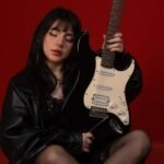 Roxie sosteniendo su guitarra en fondo rojo