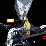 Khn de Angine de Poitrine con los brazos extendidos y su guitarra de doble mástil sobre el torso, vestido con el mameluco de lunares distintivo de la banda