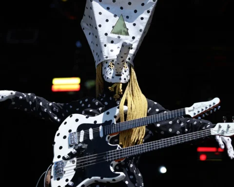 Khn de Angine de Poitrine con los brazos extendidos y su guitarra de doble mástil sobre el torso, vestido con el mameluco de lunares distintivo de la banda