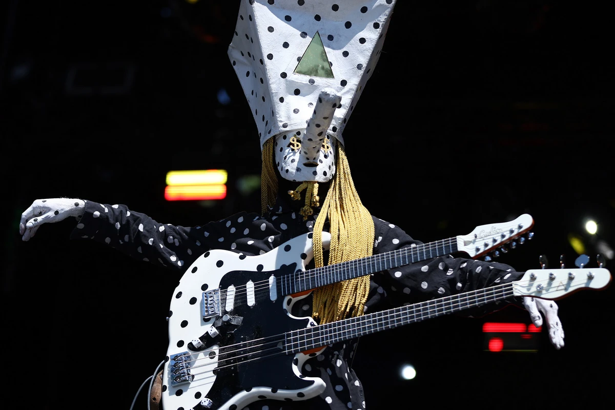 Khn de Angine de Poitrine con los brazos extendidos y su guitarra de doble mástil sobre el torso, vestido con el mameluco de lunares distintivo de la banda