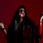 Alexander Goché con una capucha negra y las manos manchadas de negro, sobre un fondo rojo