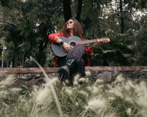 Loshe sentada sobre un muro de piedra con una guitarra, entre árboles y pasto