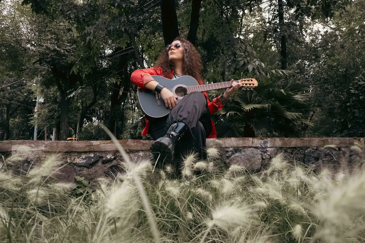 Loshe sentada sobre un muro de piedra con una guitarra, entre árboles y pasto