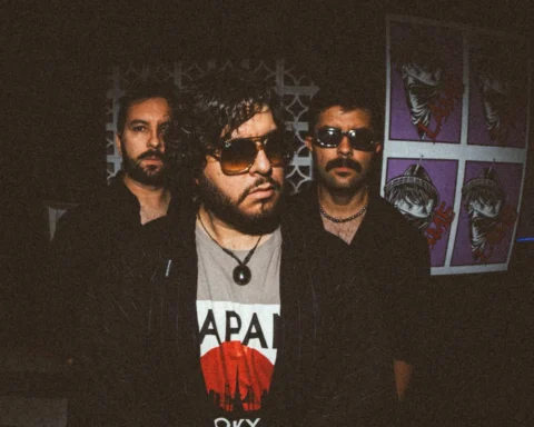 OkMark posa de pie en un espacio oscuro frente a un afiche morado: Marcos Vázquez al centro, con gafas de aviador, flanqueado por dos integrantes de la banda.