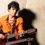 Pedro Suárez-Vértiz tocando una guitarra de doble mástil, apoyado contra un muro