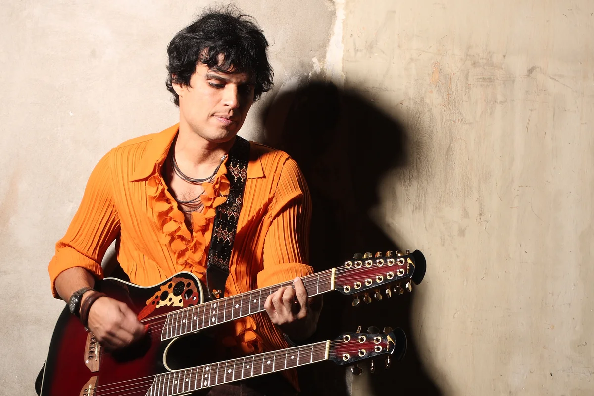 Pedro Suárez-Vértiz tocando una guitarra de doble mástil, apoyado contra un muro