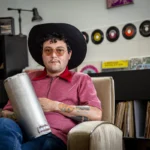 Rudy De Anda, sentado con un güiro y sombrero, en una sala con discos de vinilo