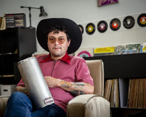 Rudy De Anda, sentado con un güiro y sombrero, en una sala con discos de vinilo