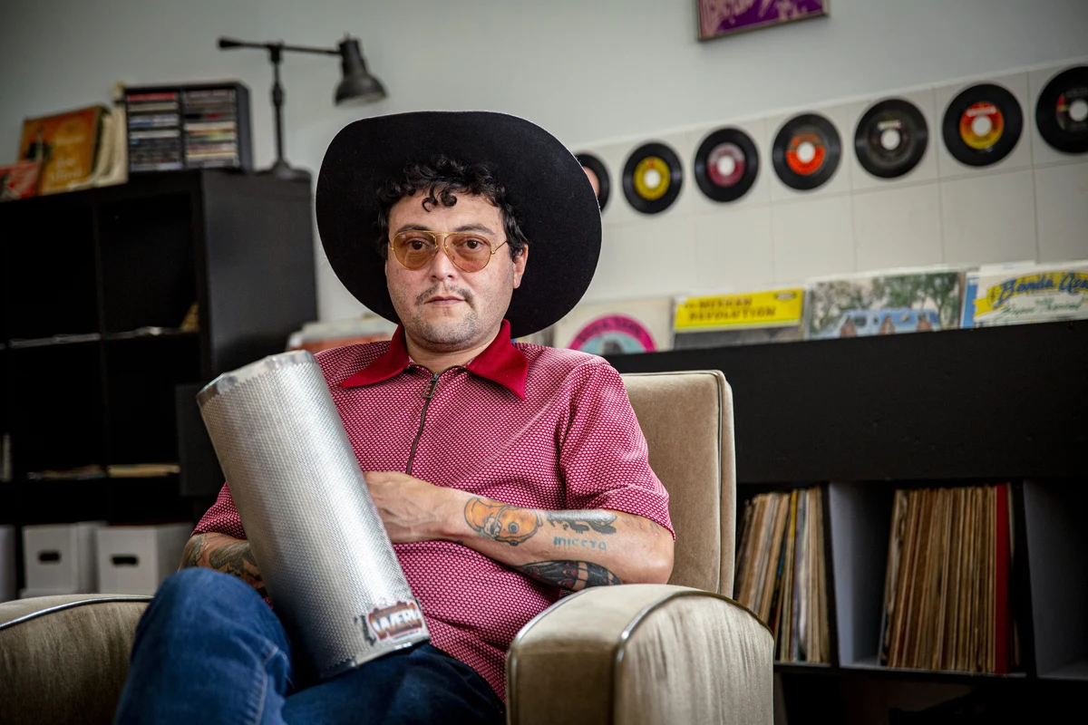 Rudy De Anda, sentado con un güiro y sombrero, en una sala con discos de vinilo